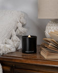 Black candle labeled 'REMONTANT' on a wooden surface with a textured white blanket and lamp in the background.