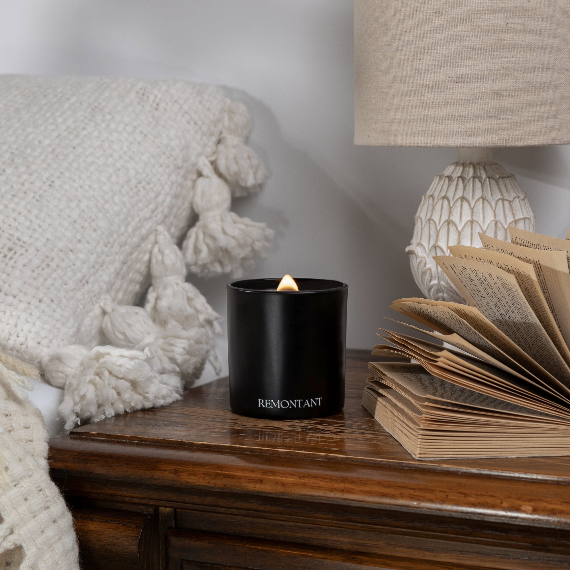 Black candle labeled 'REMONTANT' on a wooden surface with a textured white blanket and lamp in the background.